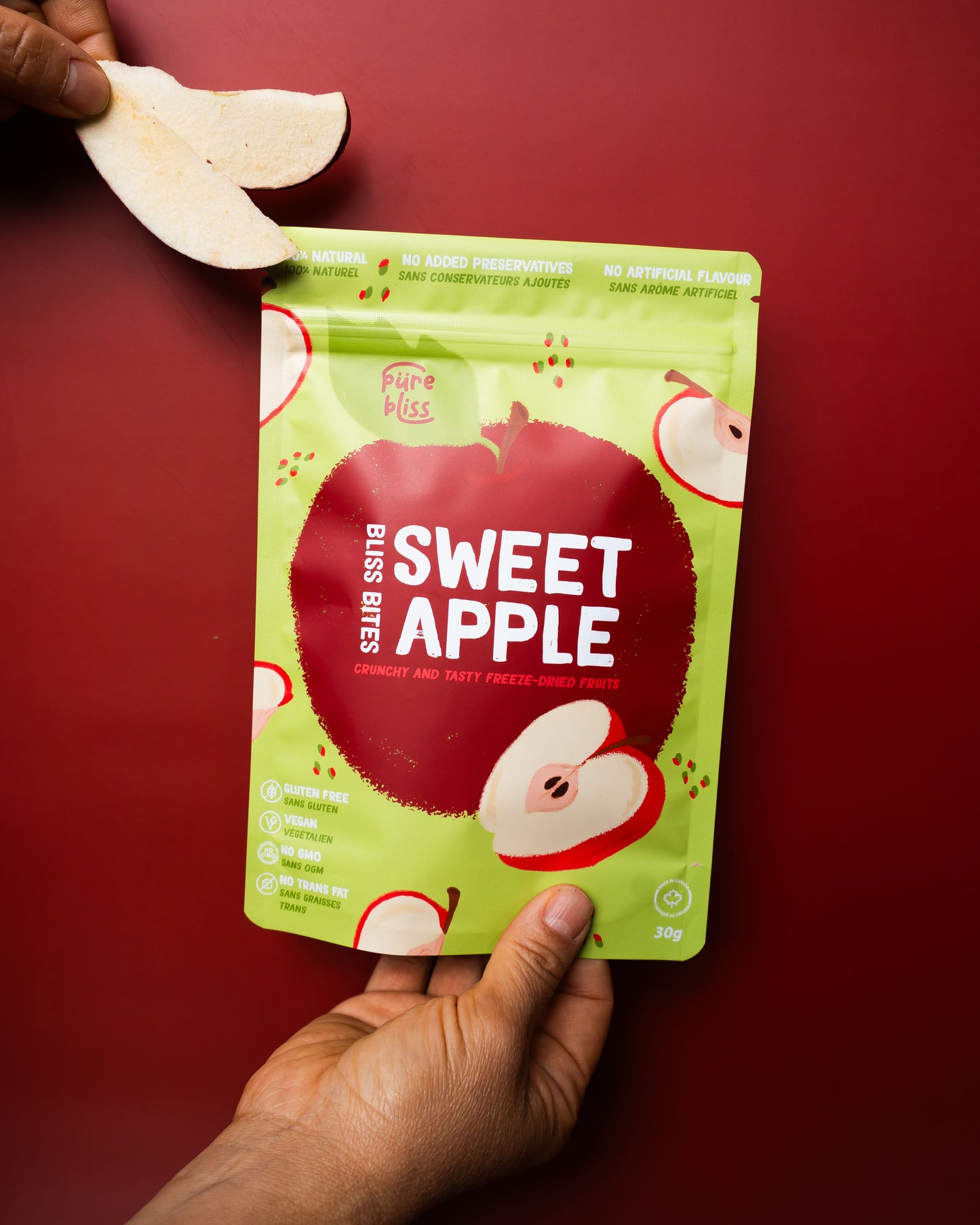 Hand holding a freeze dried apple slice and another hand holding a snack package against a red background