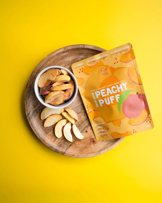 Peachy Puff snack package on a wooden plate with sliced peaches and a bowl of freeze dried fruit against a yellow background