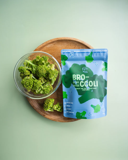 Freeze Dried broccoli in a glass bowl on a wooden plate with a blue package labeled 'Broccoli bliss bites' on a light green background.