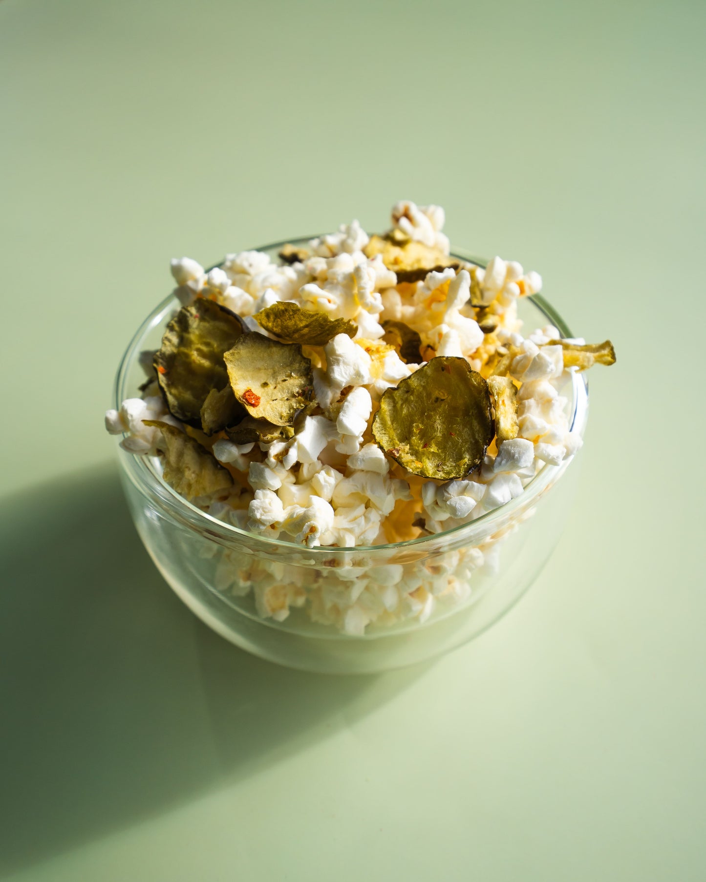 Glass bowl filled with popcorn and pickles on a light green background