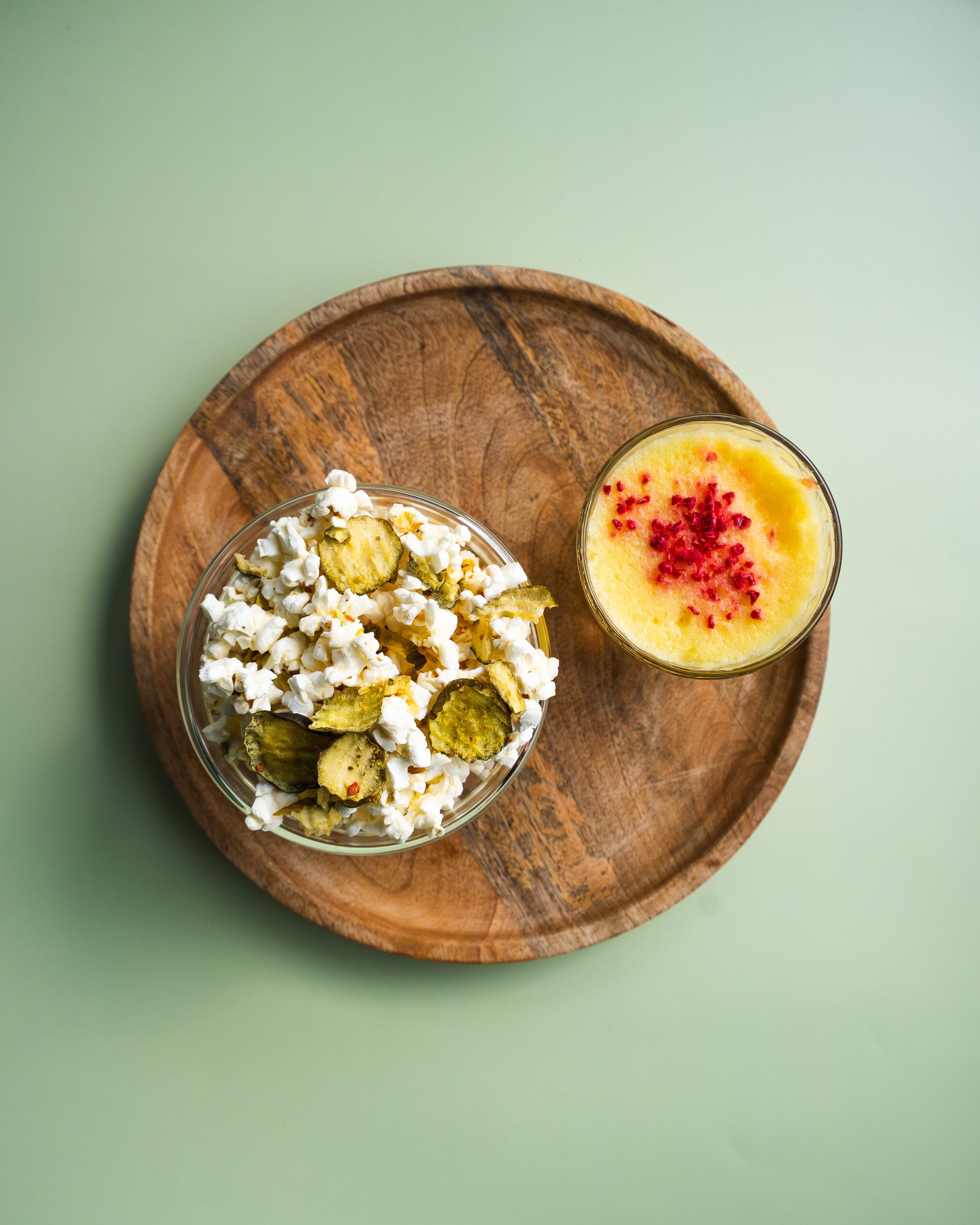 Wooden plate with a bowl of pickles and popcorn, and a glass of yellow drink with red berries on a green background