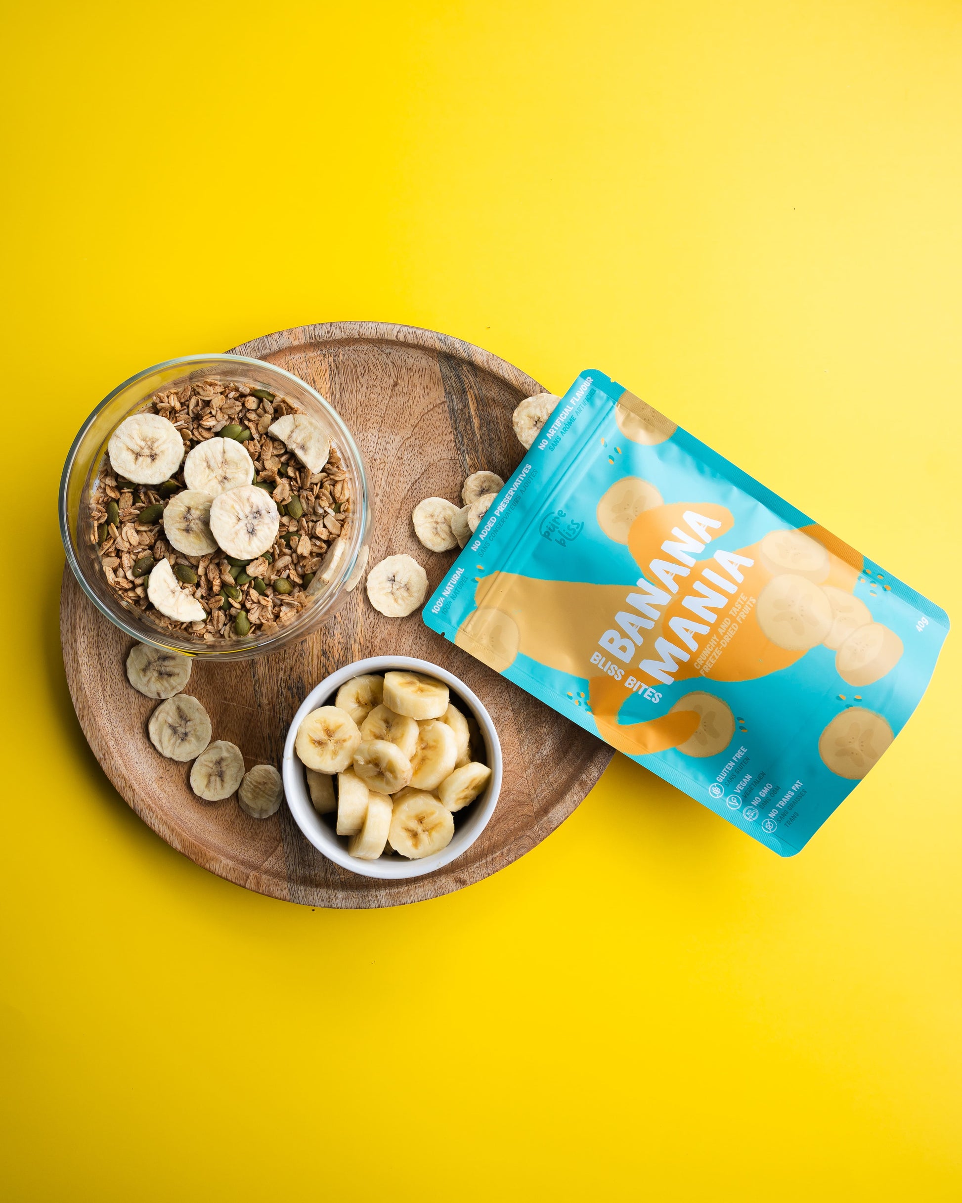 Banana freeze dried chips packaging with a wooden tray of banana slices and granola on a yellow background