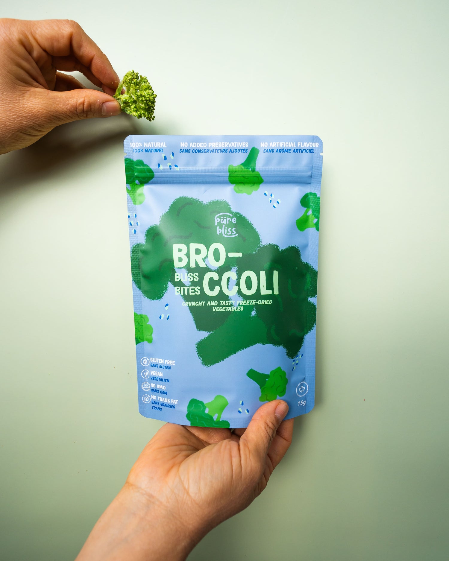 a hand holding a broccoli freeze dried bag, and another hand holding a freeze dried broccoli