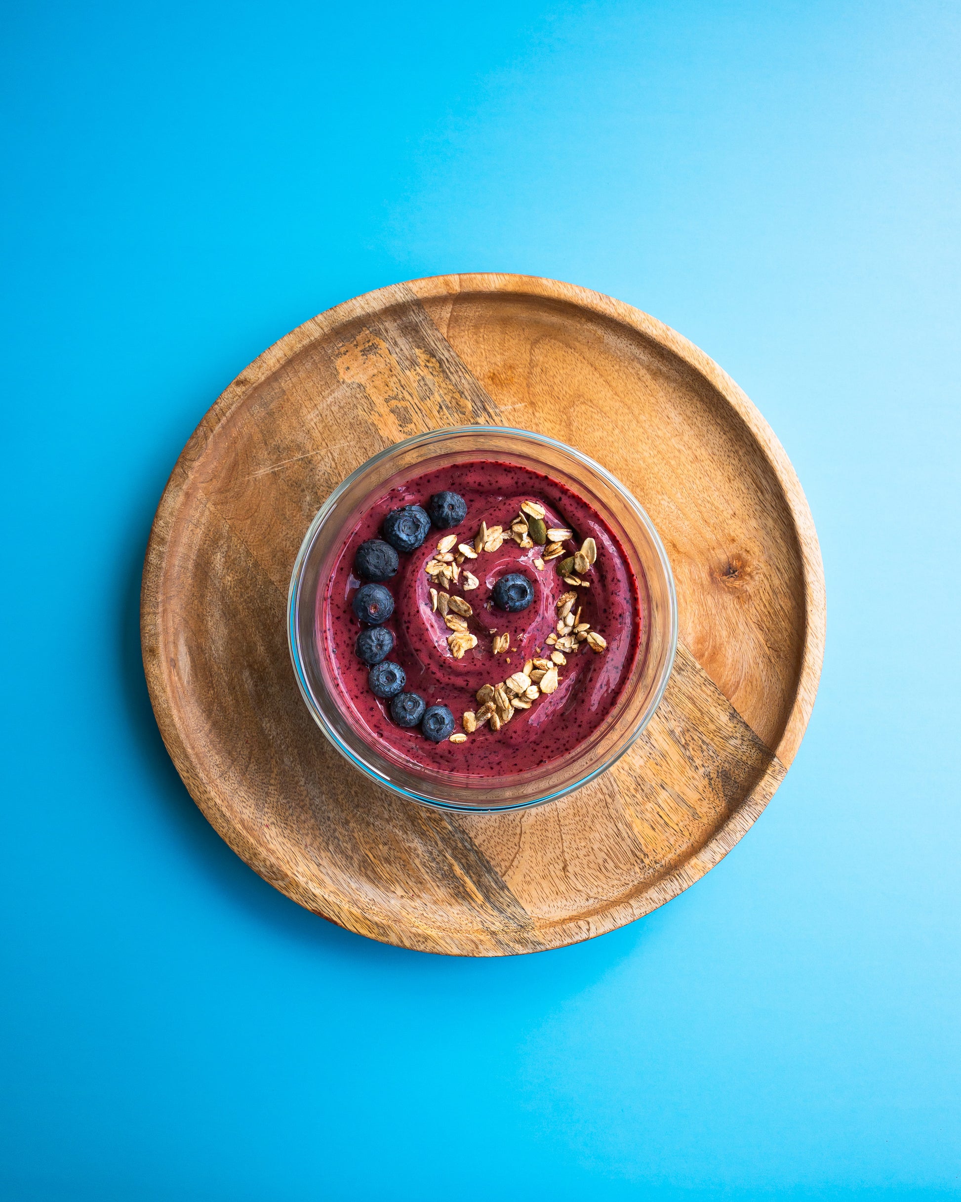 Glass bowl of berry smoothie and blueberries on a wooden plate against a blue background