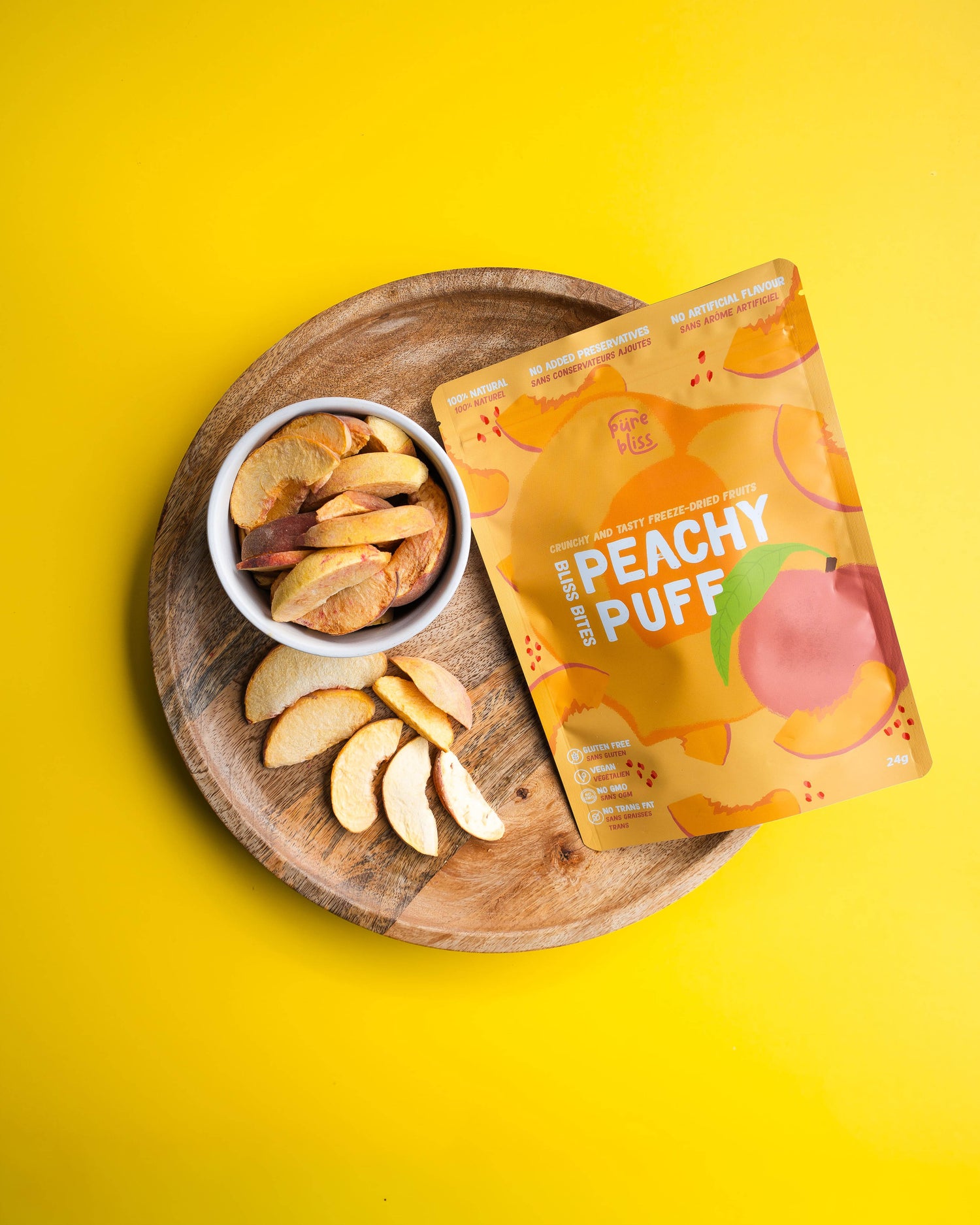 Peachy Puff snack package on a wooden plate with sliced peaches and a bowl of freeze dried fruit against a yellow background