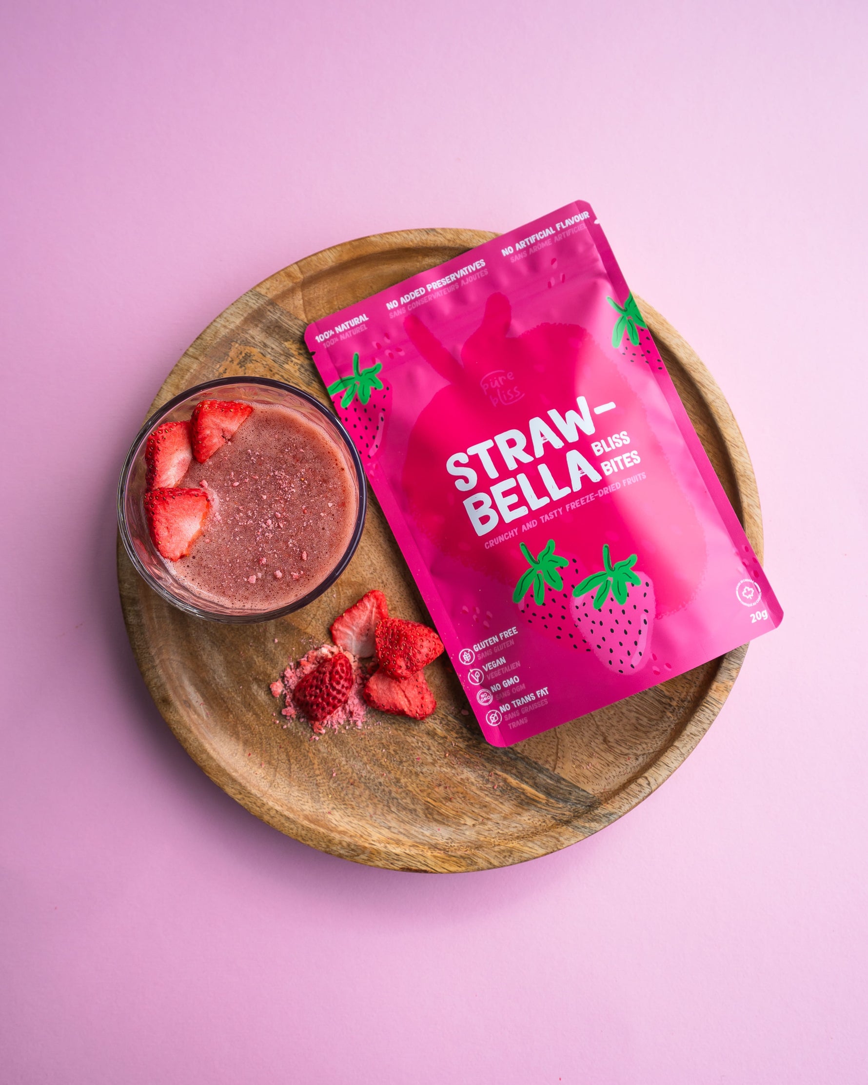 Package of StrawBella on a wooden board beside a smoothie and strawberries on a pink background