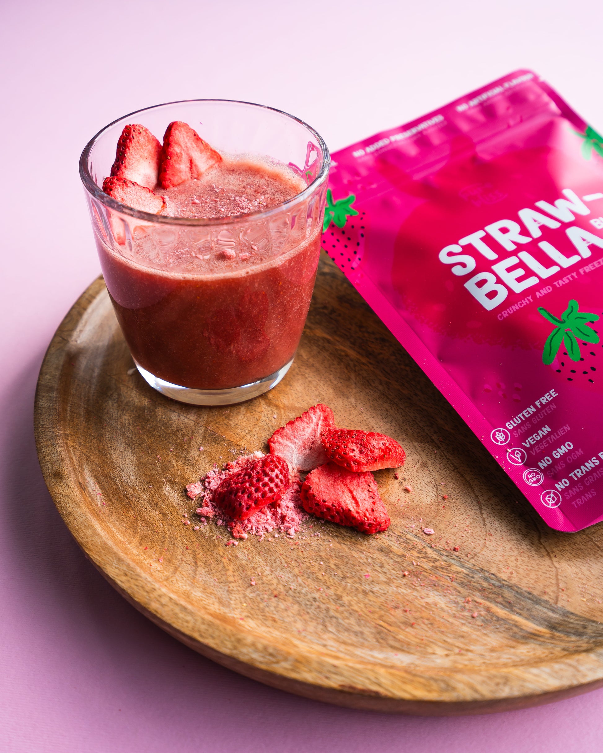 Glass of strawberry smoothie with a pink 'Straw Bella' package on a wooden tray.