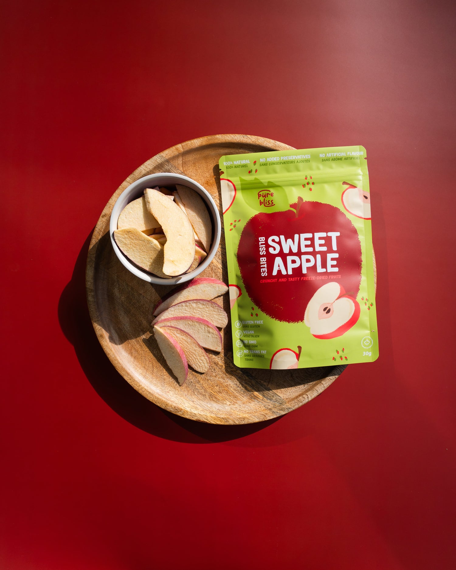 Green package of 'Sweet Apple' bites on a wooden plate with apple slices against a red background