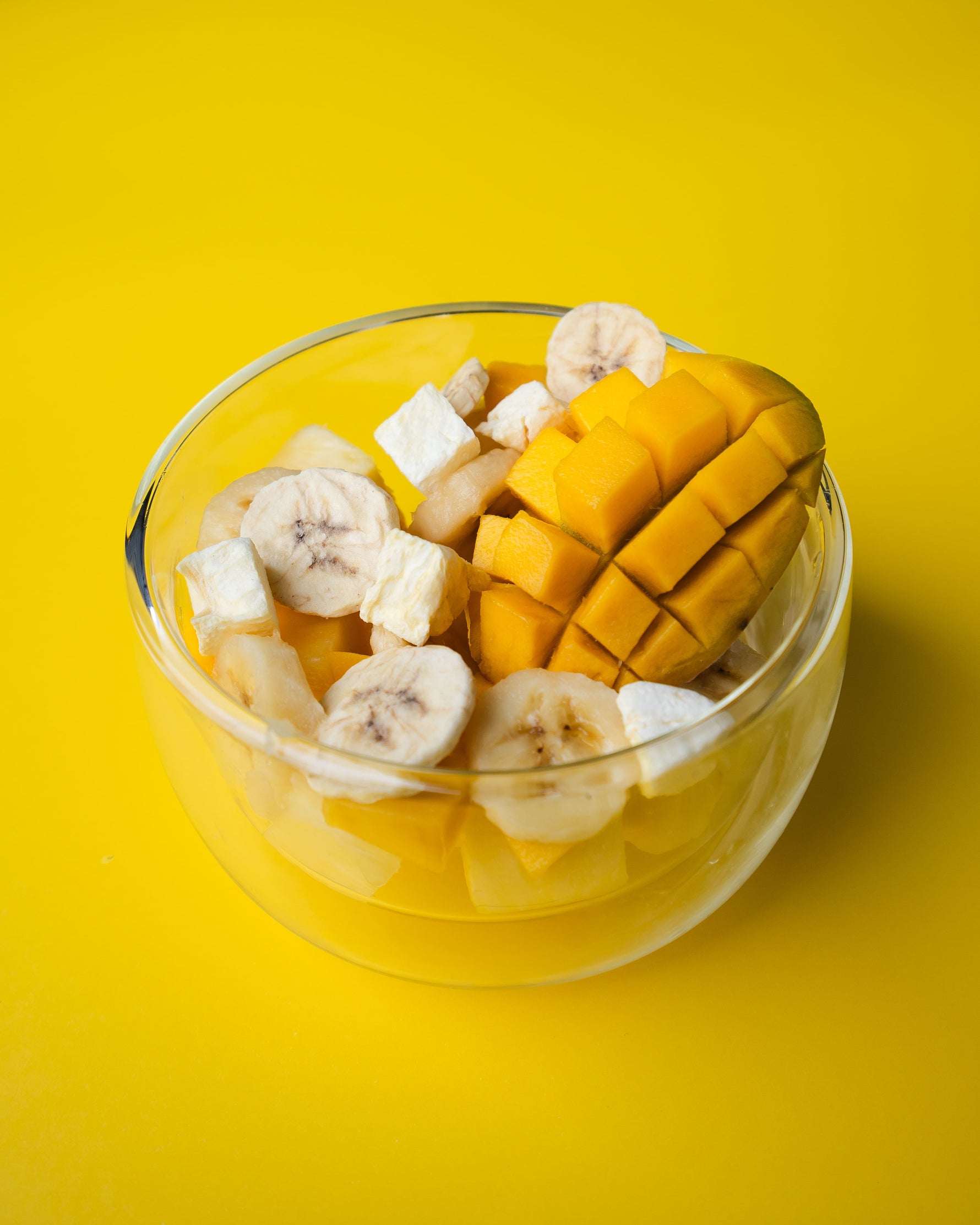 Glass bowl with sliced mango and banana and pineapple on a yellow background