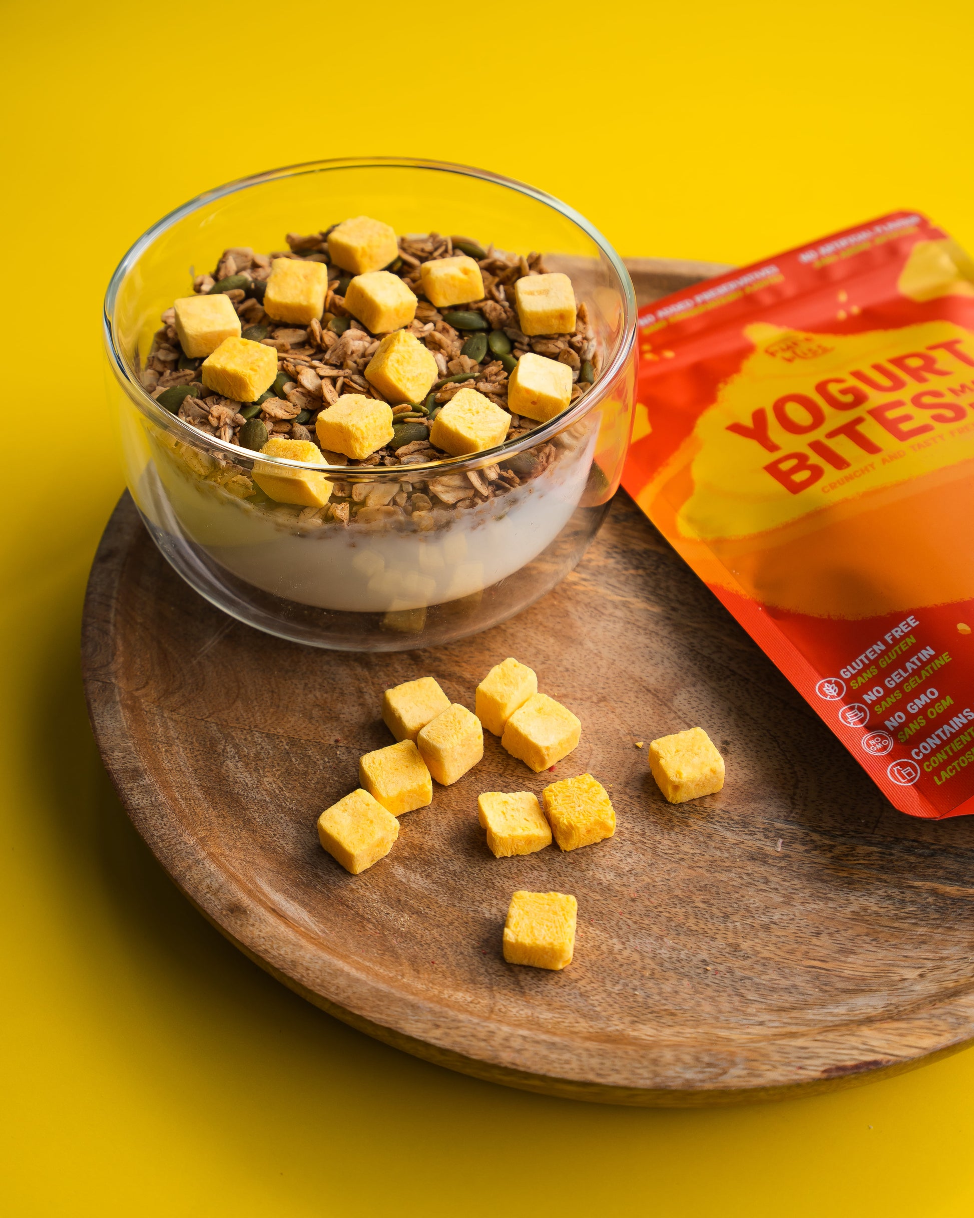 Yogurt bites in a glass bowl with a wooden tray and packaging on a yellow background