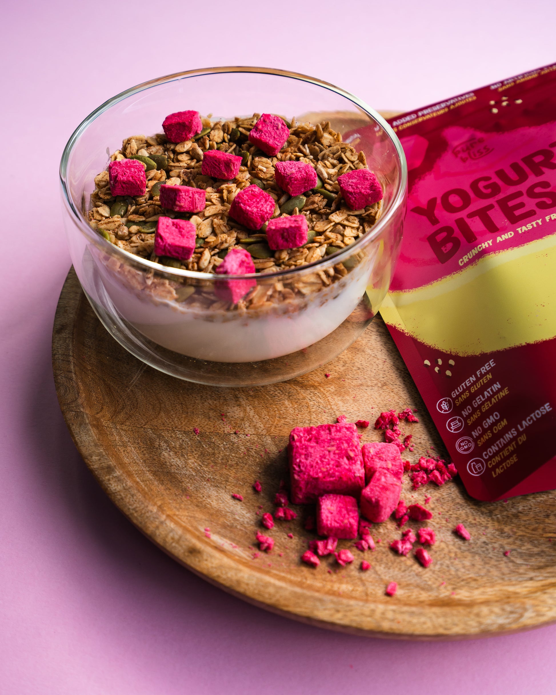 Glass bowl with granola and pink yogurt bites on a wooden plate against a pink background