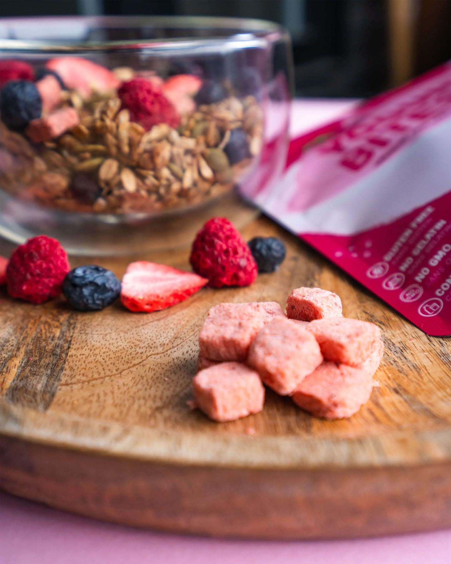 Wooden cutting board with dried fruit, a bowl of granola, and a pink package on a pink background.