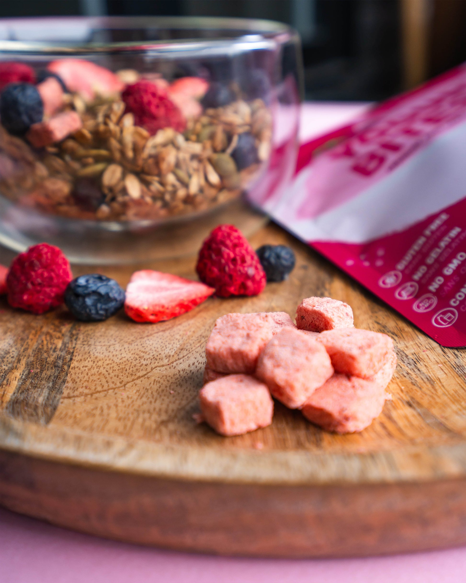 Wooden cutting board with dried fruit, a bowl of granola, and a pink package on a pink background.